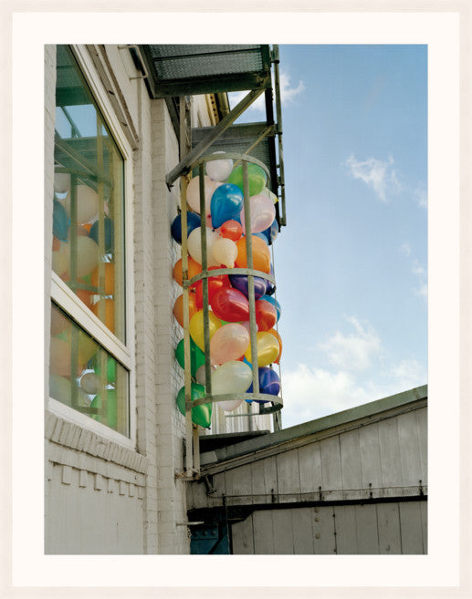 A colourful image of balloons alongside a building, by Polish artist Oliver Schwarzwald. The image is presented in a white wooden frame. 