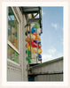 A colourful image of balloons alongside a building, by Polish artist Oliver Schwarzwald. The image is presented in a white wooden frame. 