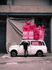 A image of a Mini car with pink presents piled on top and a woman leaning inside, by Polish artist Oliver Schwarzwald.