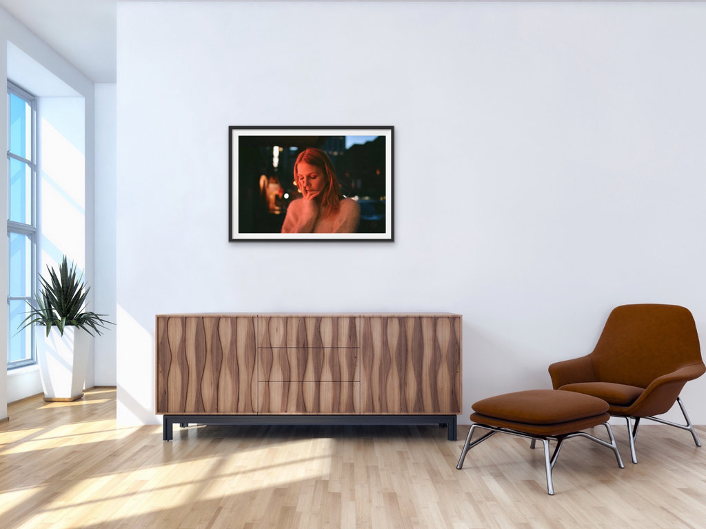 A room shot of Lost in Neon by Jenny Woods, installed above a wooden sideboard with an Eames style chair to the right. The image shows a young girl with her head in her hands, beautifully lit. 